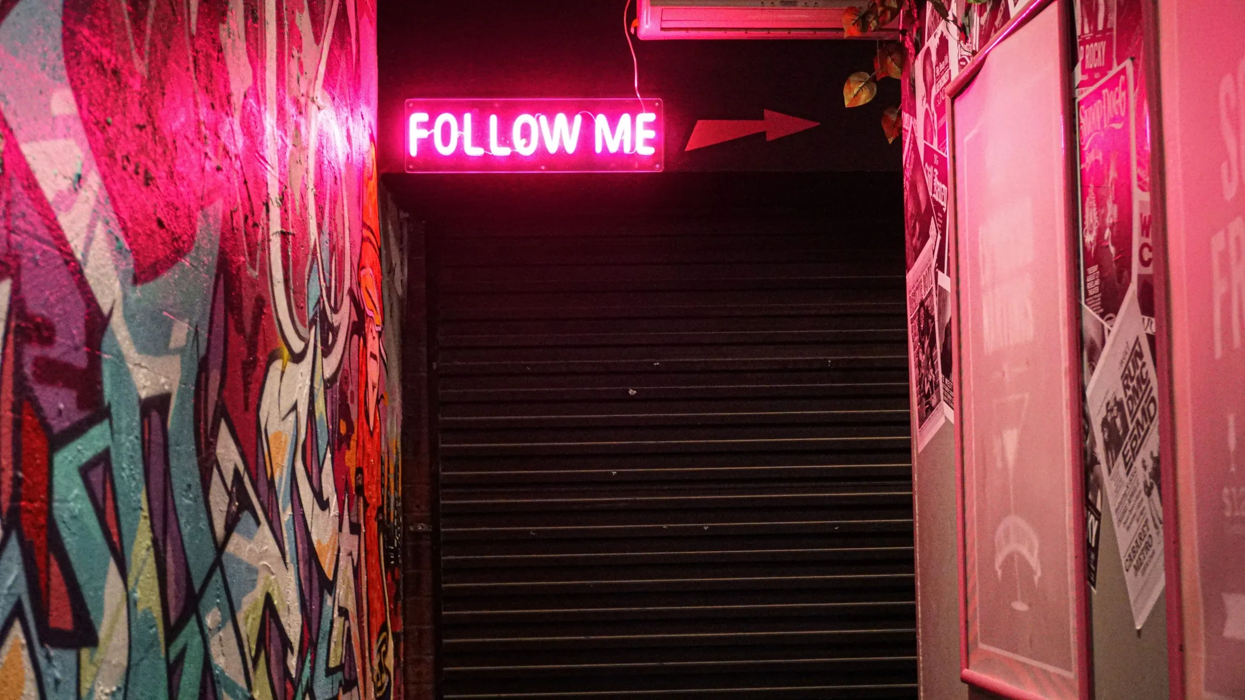 Pink neon FOLLOW ME sign above a graffitied stairwell